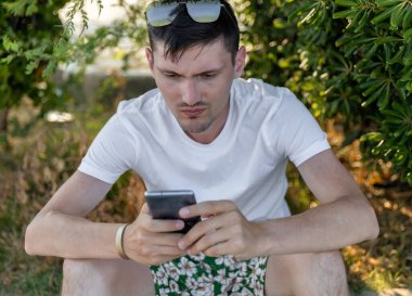 Kafasında güneş gözlüğü olan beyaz bir gencin portresi güneşli bir yaz gününde, kaşları çatık bir şekilde akıllı bir telefondan haberleşen şehir parkındaki çalıların yanındaki kaldırımda oturur.
