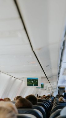 View of an electronic board with a flight path, inside a flying airplane with unrecognizable people sitting with their backs in chairs, close-up side view.