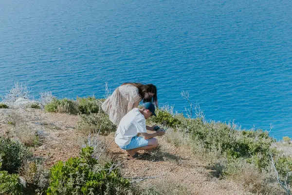 stock image Young Caucasian people, a guy and a girl, are watching a video they shot on a handheld phone on the top of a mountain against the backdrop of a blurred sea, close-up side view.