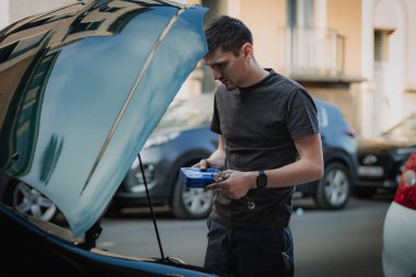 Kafkasyalı genç bir adam elinde aletlerle mavi bir kutu tutuyor ve düşünceli bir şekilde kenarda duruyor. Bir yaz günü, akşam üstü, kapalı bir şekilde bir arabanın kaputuna bakıyor.