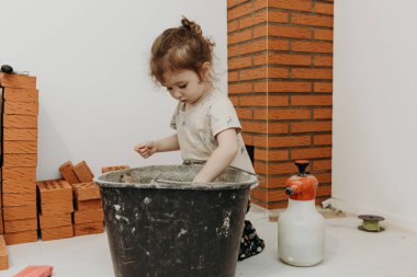 Küçük, güzel bir beyaz kız dizlerinin üstüne çöküp, tuğladan yapılmış bir şöminenin onarımı ve kurulumunun yapıldığı bir odada spatulayla bir kovada çimento harcı karıştırıyor.