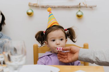 İki at kuyruklu ve kafasında konik şapkası olan güzel bir beyaz kız festival masasında bir sandalyeye oturur ve kağıt bir boruya ıslık çalar ve babası da ona eliyle yardım eder.