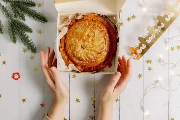 stock image Caucasian female hands clasp one royal galette in an open box with a golden crown, fir branches, a burning garland, a surprise gift and scattered confetti stars on a white wooden table, flat lay close