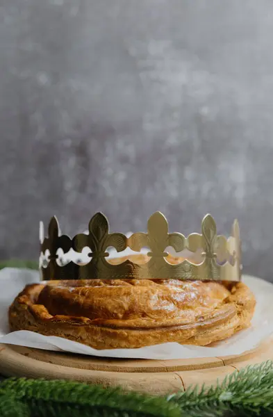 stock image One whole freshly baked king galette with golden crown on a round wooden cutting board with baking paper stands on the table with a blurred dark gray background with copy space from above, side view