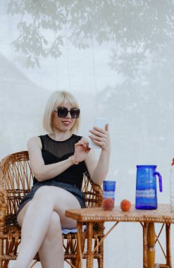 Güneş gözlüklü genç sarışın bir kızın portresi akıllı bir telefonla konuşuyor, bir ağacın altında hasır bir sandalyede oturuyor, güneşli bir yaz gününde, bir köy bahçesinde, kapalı bir köşede, perdesi olan bir ağacın altında.