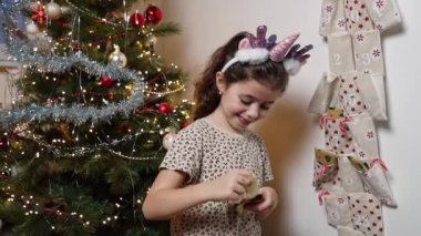 Dişsiz bir gülüşü olan güzel beyaz bir kız duvar takviminin cebinden hediyesini açar ve onu mutlu bir şekilde gösterir. Zarif bir Noel ağacının arka planında, yakın planda otururken.