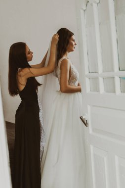 Güzel, genç bir nedime, geline kapının arkasındaki odada dikilirken beyaz bir duvak takmasına yardım eder..