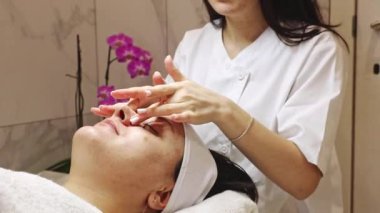 Genç, beyaz tenli bir kozmetik uzmanı burnuna nemlendirici krem sürüyor. Nazikçe masaj yapıyor ve gözleri kapalı uzanan yetişkin bir müşterinin tenine parmaklarını sürtüyor.