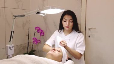 Genç, güzel, beyaz tenli, kozmetik uzmanı bir kız gözlerini bir ev güzelliğinde masaj masasına kapamış yetişkin bir müşterinin kafasına beyaz bir bandaj yapıştırıyor.