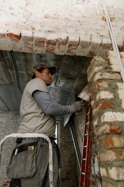 Genç bir Kafkasyalı spatula kullanarak tuğlaların üzerine taze çimento serpiştiriyor, onu düzleştiriyor ve gün boyunca eski bir evin merdiveni üzerinde dikiliyor, aşağıdan yakın çekim görüntüsü..