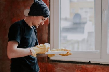 Siyah elbiseli, şapkalı, sarı eldivenli, beyaz bir genç pencerenin kenarında dikilirken pencereleri siler..