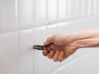 Genç bir beyaz adam, bir eliyle eski bir vidayı çıkartıyor. Banyodaki fayanslı bir duvardan küçük bir pense kullanıyor..