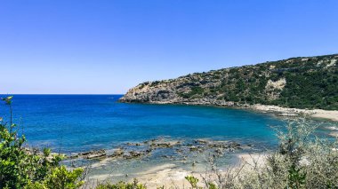 Güneşli bir yaz gününde Yunanistan 'ın Rodos adasındaki Ege Denizi' nin sularından, dağlardan ve bitki örtüsünden sarkan kayalarla ıssız bir sahil manzarası.