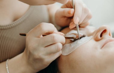 Genç, güzel, beyaz bir kozmetik uzmanı kızın portresi. Ofisindeki kozmetoloji masasında uzanan bir kadın müşteriye cımbızla sağ gözüne doğal kirpiklerini serer.