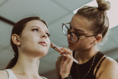 Genç, yakışıklı bir beyaz makyaj sanatçısı, sabahın erken saatlerinde bir güzellik salonunda, aşağıdan yakın çekim manzaralı bir sandalyede oturan bir kızın elmacık kemiklerine fosforlu kalem sürüyor. Adım adım..
