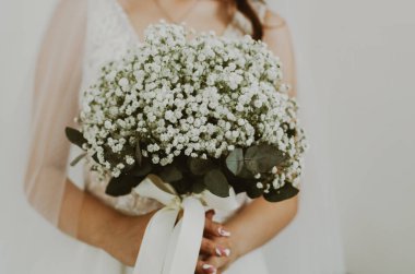 Portrait of one unrecognizable young Caucasian bride in a white dress and veil holding a wedding bouquet of white boutonnieres in her hands in front of her, side view, close-up. clipart