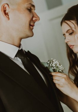 Genç ve güzel bir esmer gelinin portresi, siyah takım elbiseli damadın göğüs cebine küçük bir buket çiçek yerleştiriyor. O da bir yaz sabahı odada, ona nazikçe bakıyor. Düğün gelenekleri konsepti.