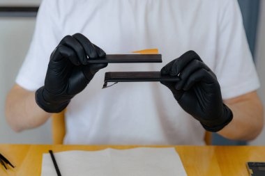 Beyaz tişörtlü, siyah lateks eldivenli, tanınmayan genç bir beyaz adam, bir akıllı telefon için eski şişmiş ve yeni pilleri gösteriyor. Bir yaz günü, yakın planda, mutfakta bir masada oturuyor.