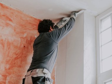 Üniformalı, kıvırcık saçlı, genç bir beyaz erkek alçı, pencerenin yanındaki duvardan büyük bir spatula ve macun çıkarıyor, merdivenin alt tarafında, yakın plan.