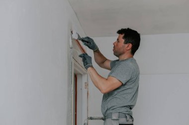 Gri üniformalı ve eldivenli genç bir beyaz tenli esmer, inşaat ağını beyaz macunla kaplıyor, kapının önünde duran bir merdiven, aşağıdan yakın çekim görüntüsü