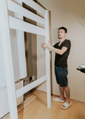 Genç bir beyaz adam yerde duruyor ve bir beyaz yatağı tutuyor. Akşam, odada. Yan tarafı seçici bir odak ile yakın plan..