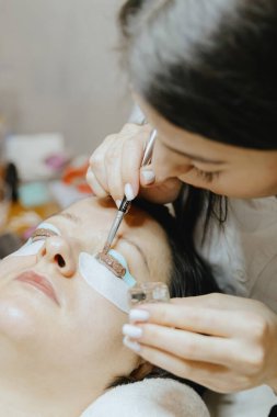 Beyaz önlüklü, yarı tanınmış beyaz bir kozmetik uzmanı kızın portresi sol gözünün silikon kalıbına kahverengi kirpik boyası sürerken esmer bir kadına fırça kullanıyor.