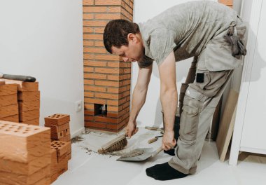 Genç bir beyaz erkek müteahhit, bir süpürgeyle uzanan tuğlanın ortasında kurumuş çimento kalıntılarını süpürüyor ve odanın içinde bükülmüş vaziyette faraş gibi duruyor. yan görünüm kapat