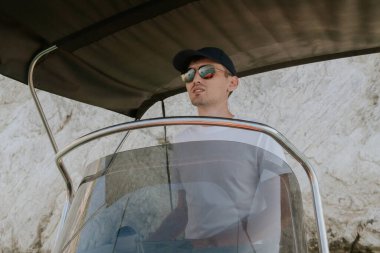 Şapkalı, güneş gözlüklü ve beyaz tişörtlü genç bir beyaz adam kayalık bir sahilden uzaklaşır. Güneşli bir yaz gününde dümeni yönetir..