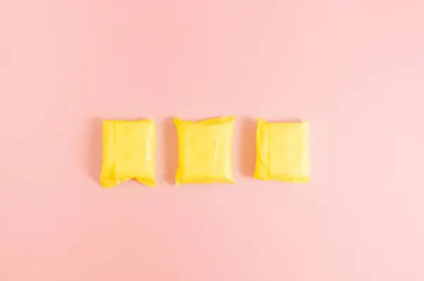 Stock image Three feminine pads in yellow packages lie in a row in the center on a pink background, flat lay close-up with copy space on the sides.