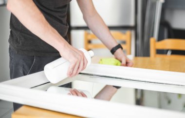 Koyu renk elbiseli, tanınmayan genç bir beyaz adam bir şişeden aynaya aseton dökerek boyadan sonra lekeleri temizler. Yazın mutfakta dikilirken yansıtır.