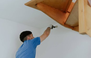 Beyaz tenli genç bir esmer, mavi bir tişörtle yan yana durur ve duvarların köşelerini tahta bir merdivenin altına beyaz boyayla boyar. Küçük bir fırça kullanır.