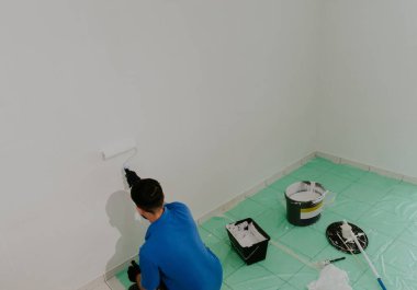 Mavi tişörtlü, koyu renk saçlı, genç bir beyaz adam dizlerinin üzerinde yeşil bir bez ile oturuyor ve yuvarlak, yakın bir üst görüşe sahip beyaz boyalı bir duvar çiziyor.