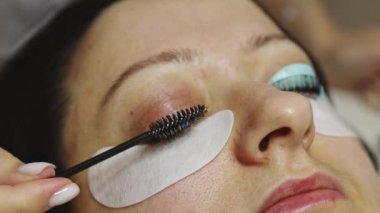 Sağ gözünün kirpiklerini siyah bir fırçayla fırçalayan beyaz, tanınmayan genç bir kız kozmetik uzmanının elleri. Ev güzellik salonundaki kozmetoloji masasında uzanmış bir müşteri.