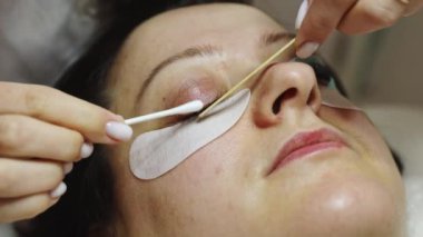 Sağ gözünün kirpiklerini ahşap ve pamuklu bezle kaplayan beyaz bir kozmetik uzmanının elleri. Bir müşteri kozmetoloji masasında yatıyor.