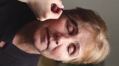 Genç bir beyaz kozmetik uzmanı, gözleri kapalı yaşlı bir kadına doğru renkli kaşlarını tarıyor. Akşamları bir evin güzellik salonunda, lambanın altında oturuyor.