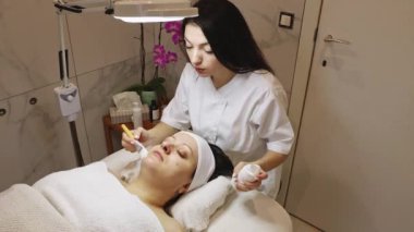 Genç bir kozmetolog kız, bir güzellik salonundaki masaj masasında gözleri kapalı yatan bir müşteriye fırça yardımıyla çenesine ve sol yanağına temizleyici köpük sürer.