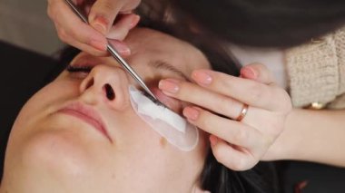 Bir müşterinin sol gözündeki doğal kirpikleri ayıran beyaz tenli genç kozmetikçi. Yapay olanları yapıştırmak için cımbız kullanıyor. Bir bahar günü ev ofisinin kozmetoloji masasında yatıyor.