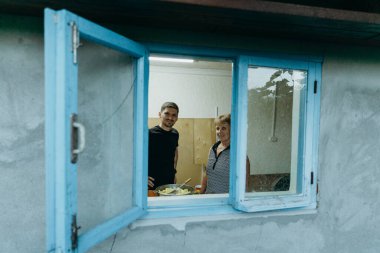 Genç bir torun ve yaşlı bir büyükanne kameraya bakıp patates kızartma tavasında patates kızartıyorlar. Yazın eski bir kır evinde, pencerenin kenarından bakıyorlar.
