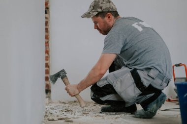 Gri tişörtlü ve şapkalı genç bir beyaz adam, kapının alt tarafındaki eski macunu baltayla temizlerken aşağıdan seçici bir odak ile çömeliyor. İnşaat işi