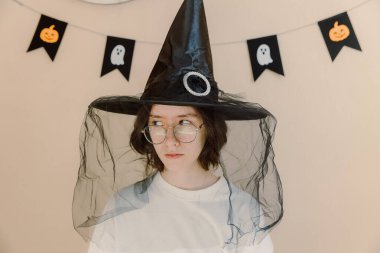 Portrait of one Caucasian beautiful happy teenage girl in a witch hat with black tulle Halloween, who looks slyly to the side, standing against the background of a wall with flag decor, preparing for clipart