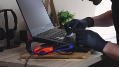 Siyah lateks eldivenli genç bir beyaz adamın elleri, bir yaz günü yatak odasındaki ofis masasında otururken dizüstü bilgisayarın klavyesindeki çıkartmaları kağıt peçeteyle temizliyor.