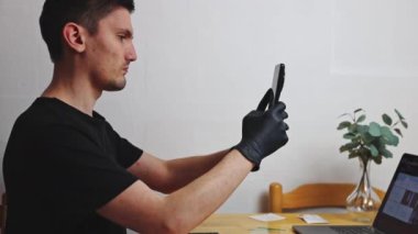 Siyah lateks eldivenli genç bir beyaz adam, koruyucu filmi tamir etmeden ve değiştirmeden önce akıllı telefonun kamerasını dikkatle inceliyor, mutfaktaki ahşap bir masada dizüstü bilgisayarla oturuyor.