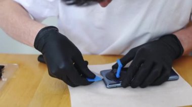 Beyaz tişörtlü ve siyah lateks eldivenli genç bir beyaz adam özel bir mavi tabak ve mutfak bıçağı kullanarak bir akıllı telefonun arka kapağını açar ve masada otururken onu tamir eder.