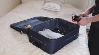 Tek elli, koyu renk elbiseli, beyaz, genç bir adam bir yaz günü yatak odasındaki eşyaların olduğu açık bir bavula dalış maskesi koyar.
