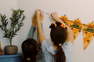 İki güzel beyaz kızın portresi mutfağı şenlikli turuncu Cadılar Bayramı bayraklarıyla süslüyor, onları çift taraflı bantlara yapıştırıyor, birbirlerine yardım ediyor ve ayakta duruyorlar.