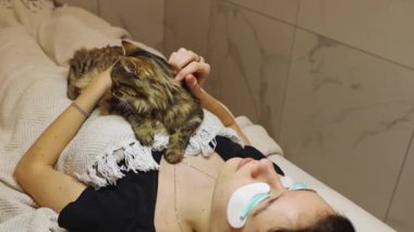 Beyaz tenli güzel bir kız karnında uyuyan bir kediyle uzanmış, kirpik kaldırma prosedürünü beklerken onu okşuyor, orta ölçekli bir yan görüş. Kirpik.