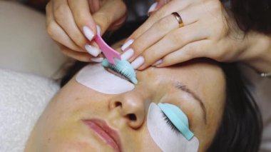 Sağ gözünün kirpiklerini özel pembe bir plastik fırça kullanarak silikon bir kalıba yapıştıran beyaz bir kozmetikçinin elleri. Bir kadın güzellik salonunda yatıyor.