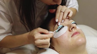 Sol gözünün kirpiklerini silikon bir formda kaplayan beyaz tenli genç bir kız pamuklu çubukla kalan kahverengi boyayı temizliyor, kozmetoloji masasında yatan bir kadın.