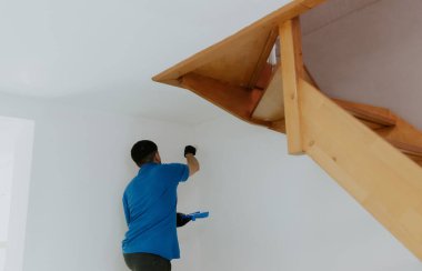 Mavi tişörtlü, yakışıklı, koyu renk saçlı beyaz bir adam yarım yamalak duruyor ve beyaz bir fırçayla duvarın köşelerini boyuyor ahşap merdivenler ile kapı arasının alt tarafını boyuyor.