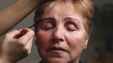 Genç bir beyaz kozmetik uzmanı, yaşlı ve memnun bir kadına doğru renkli kaşını dikkatlice tarıyor. Gözleri kapalı, akşamları ışık altında bir güzellik salonunda oturuyor.